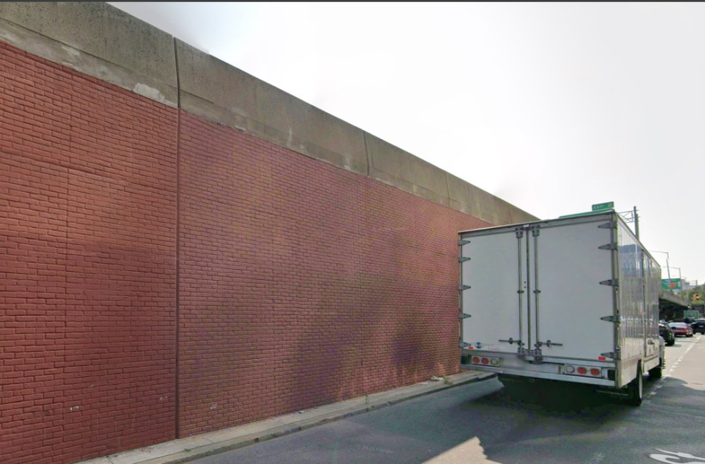 Tall brick ramp wall along the local street next to the I-87 edge, white box truck for scale.